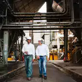 Two workers wearing safety helmets and protective gear walk through an industrial facility with large metal structures and pipes.