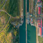 Aerial view of a ship passing through a canal lock surrounded by green vegetation, industrial buildings with red roofs, and blue water on both sides.