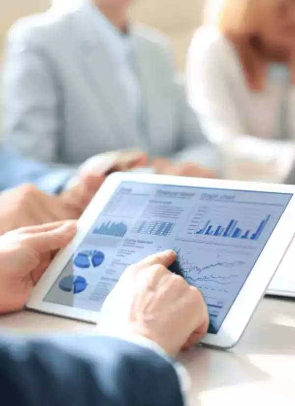 Close-up of a person using a tablet displaying financial charts and graphs during a business meeting, with colleagues in the background.