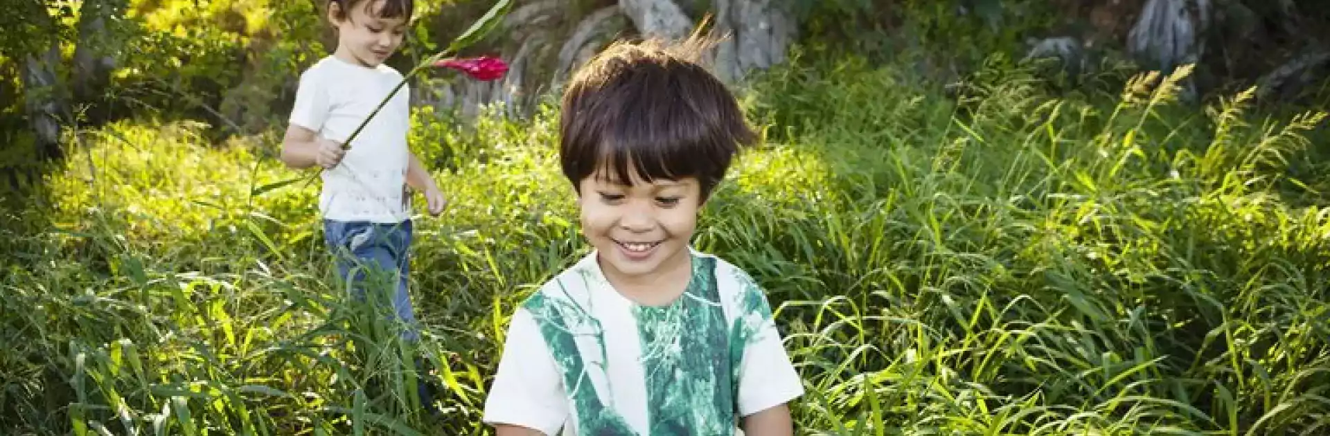 Two young children playing in tall green grass outdoors. One child in the foreground is smiling and looking down, while the other child in the background holds a red flower. Sunlight filters through the trees, creating a cheerful and natural setting.