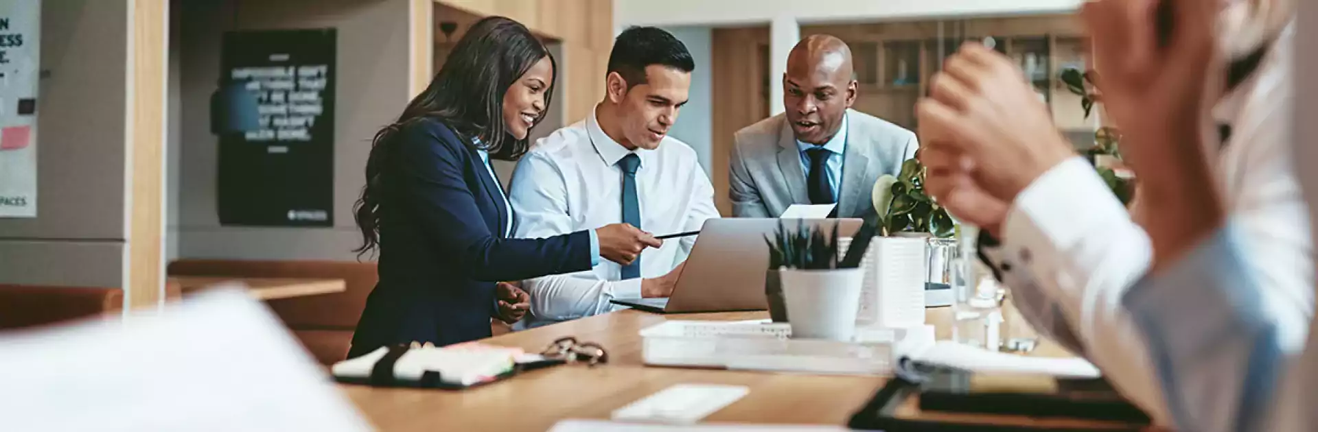 Three business professionals collaborating at a modern office table, discussing documents and working on a laptop, with two colleagues blurred in the foreground. The workspace features wooden decor and pendant lighting.