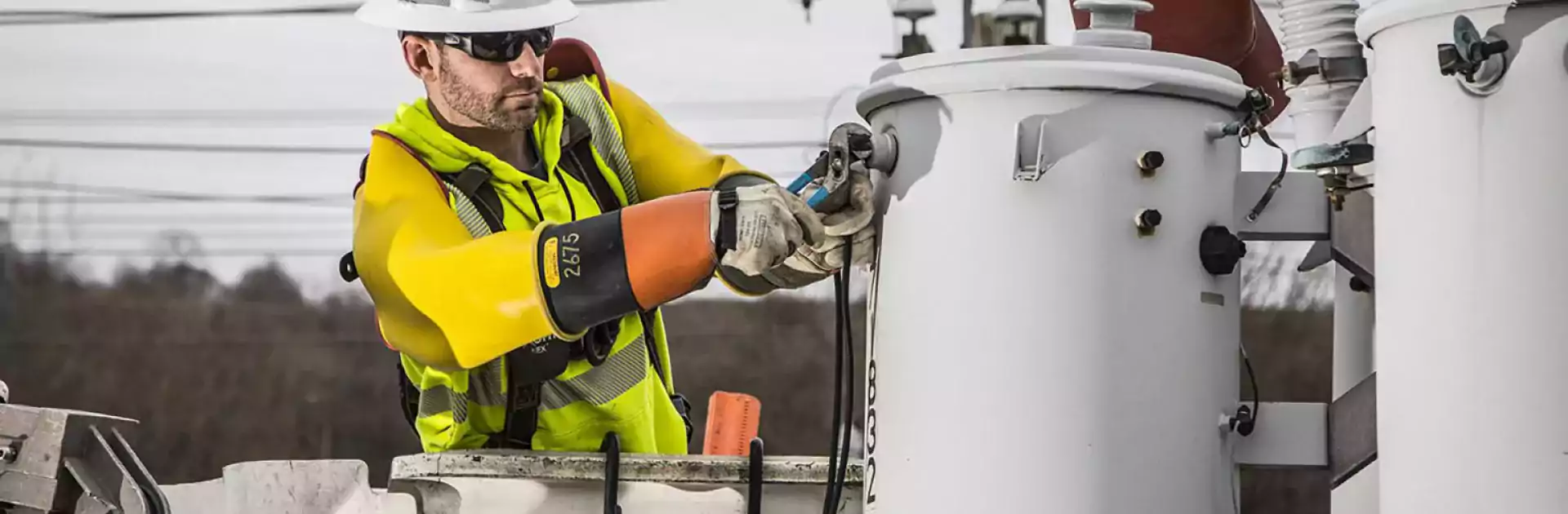 Utility worker wearing a hard hat, safety glasses, and insulated gloves repairs electrical equipment on a power pole using pliers while standing in a bucket truck. The worker is surrounded by transformers and power lines, demonstrating electrical safety procedures.