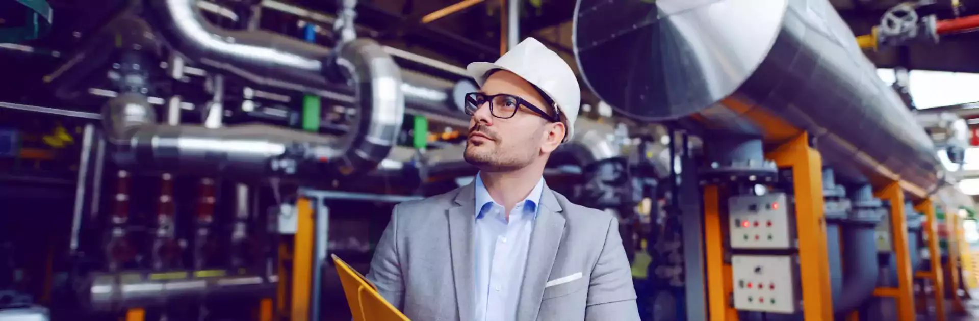 A man wearing a suit, glasses, and a white hard hat stands in an industrial facility holding a yellow folder. He is inspecting the machinery and large metal pipes in the background, suggesting he is conducting a safety or operational inspection.