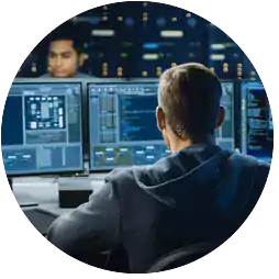 Person sitting at a desk with multiple computer monitors displaying code and data in a control room, with another person visible in the background.