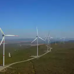 A row of wind turbines stands on a green landscape under a clear blue sky, generating renewable energy. The turbines are aligned along a winding road, surrounded by vegetation, with distant hills visible in the background.