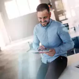 Smiling man in a light blue shirt sitting on a desk in a modern office, using a digital tablet. The background is bright with large windows and office furniture.