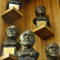 Five bronze busts of men are displayed on wooden shelves mounted on a wood-paneled wall. Each bust has a plaque at its base, and the arrangement creates a sense of depth and prominence for each sculpture.