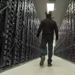 A person wearing a helmet walks down an aisle between rows of large electronic equipment racks in a modern data center, viewed from behind.