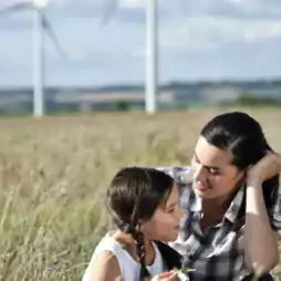 A woman and a young girl sit together in a grassy field, smiling and talking, with wind turbines in the background under a partly cloudy sky.
