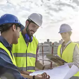Three construction workers wearing safety vests and helmets review blueprints and take notes at a building site under a partly cloudy sky.