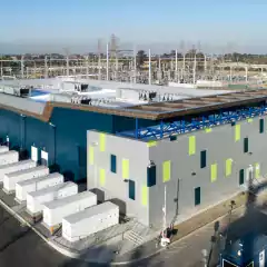 Aerial view of a modern energy storage facility with a large rectangular building, multiple white battery storage units outside, and electrical infrastructure including power lines and transformers in the background.