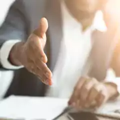 A businessperson wearing a suit extends their hand for a handshake, with documents and a smartphone visible on the table in a bright office setting.