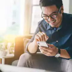 Smiling man wearing glasses and a blue shirt sitting on a couch, using a smartphone in a bright living room with sunlight coming through the window.