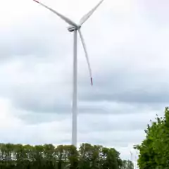 A tall wind turbine stands next to a dirt road with green trees and grass under a cloudy sky. Additional wind turbines are visible in the background, indicating a wind farm in a rural landscape.