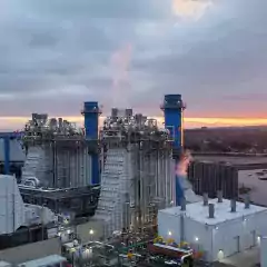 Industrial power plant with large blue chimneys and complex piping system, emitting steam at sunset. The facility is surrounded by urban landscape and water, with a dramatic cloudy sky and orange horizon in the background.
