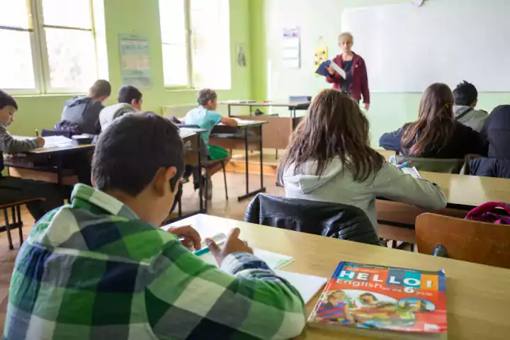Students sit at desks in a classroom, writing in notebooks while a teacher stands at the whiteboard giving a lesson. An English textbook is visible on a desk in the foreground. The classroom is well-lit with natural light from large windows.