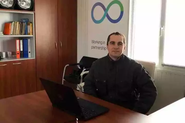 Man sitting at a desk with a laptop in an office, next to a window. Behind him is a cabinet with books and safety helmets, and a banner with the aes logo and the text 'Working in partnership.' The office is well-lit with natural light.