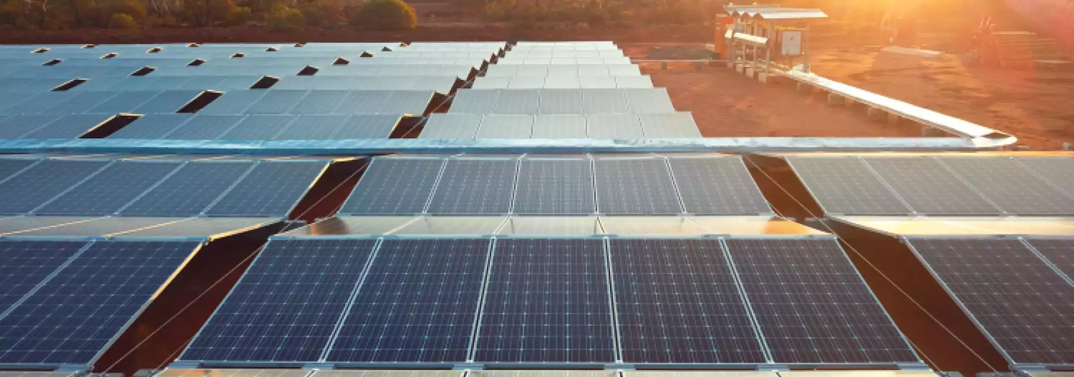 Rows of solar panels installed in a large open field with sunlight shining in the background, surrounded by trees and a small utility structure.