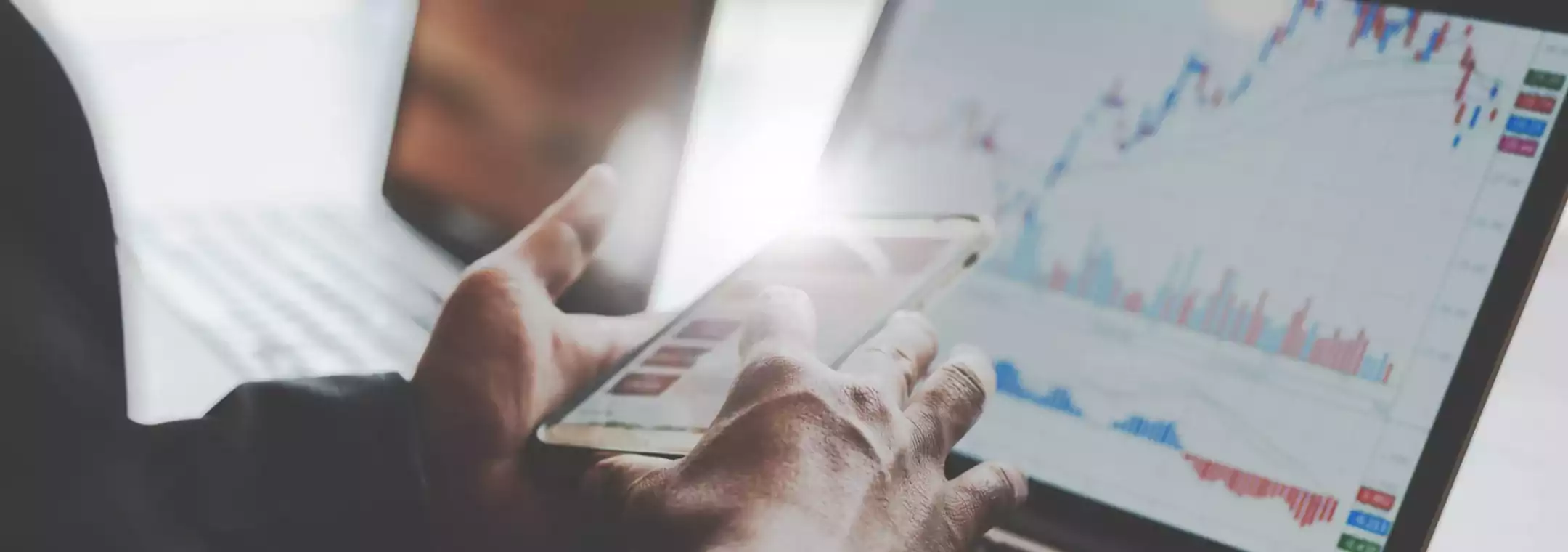 Close-up of a person using a smartphone and laptop with financial charts and graphs displayed on the screen, indicating stock market analysis or trading activity.