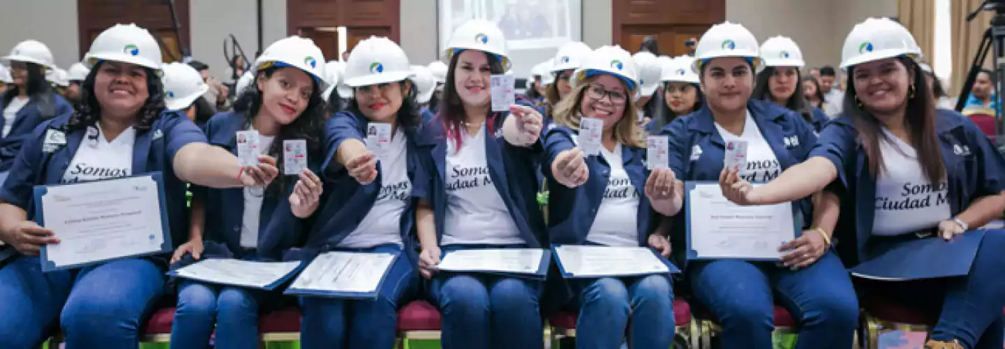 A group of women wearing white construction helmets and blue jackets sit in a row, holding certificates and ID cards, smiling at the camera in a formal indoor setting.