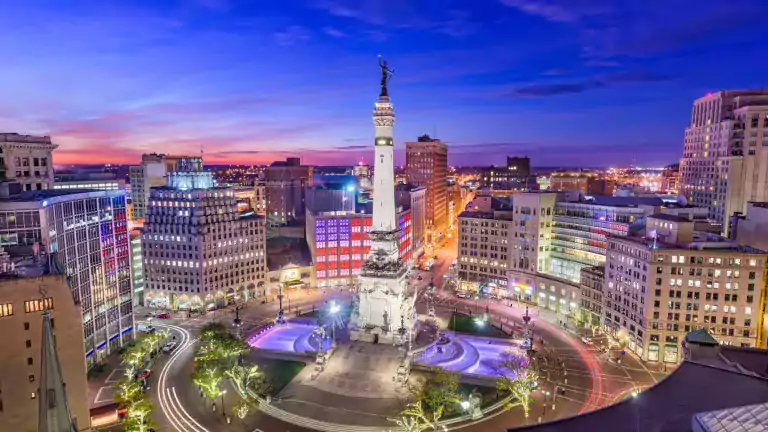 Aerial view of downtown Indianapolis at dusk, featuring the illuminated Soldiers and Sailors Monument surrounded by city buildings, colorful lights, and light trails from vehicles.
