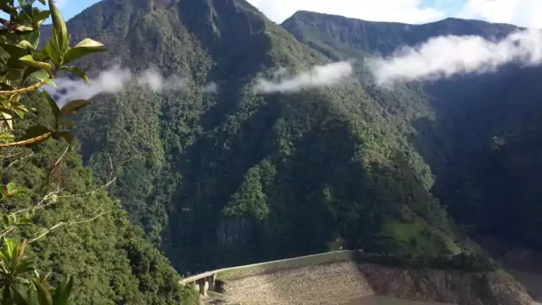 A lush green mountain landscape with a dam at the base, a winding river, and low clouds drifting across the slopes. Dense vegetation covers the mountains, and some leaves are visible in the foreground.