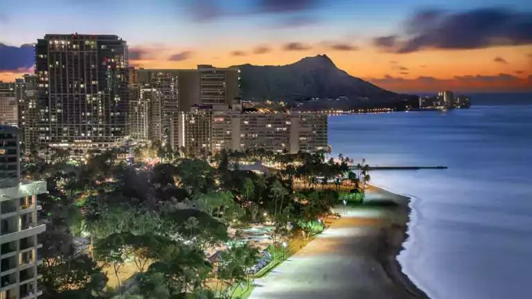 A scenic view of Waikiki Beach in Honolulu at sunset, featuring illuminated high-rise hotels, palm trees, and the iconic Diamond Head mountain in the background, with calm ocean waves along the shoreline.