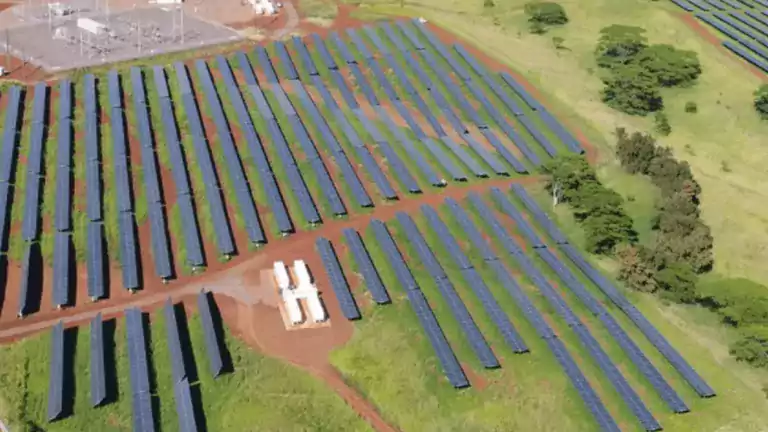 Aerial view of a large solar farm with rows of solar panels installed on green grass and red soil, surrounded by trees and a substation in the background.