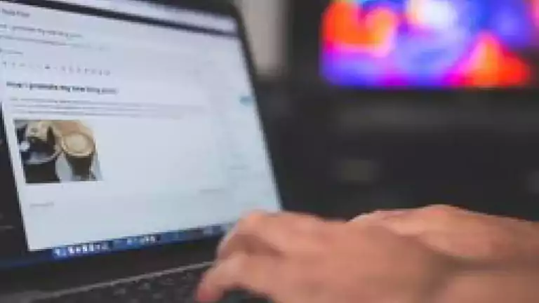 Person typing on a laptop with a blog post editor open on the screen, showing a photo of coffee. A colorful, blurred television is visible in the background.