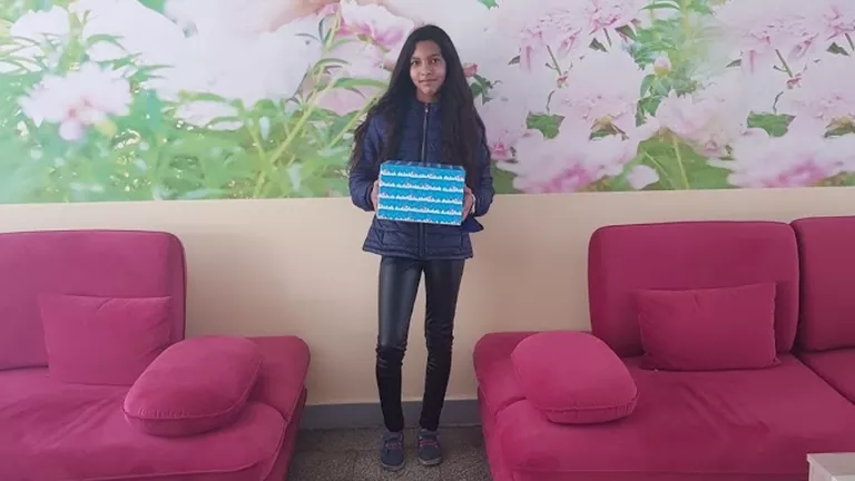 A girl with long dark hair stands between two pink sofas, holding a blue gift box with white patterns. She is wearing a dark jacket and black pants. The background features a large floral mural with pink flowers and green leaves.