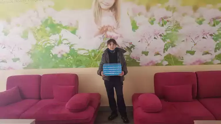 A child wearing a jacket and dark pants stands indoors holding a blue box, positioned between two pink sofas. Behind the child is a large mural of a girl surrounded by pink flowers, creating a cheerful and inviting atmosphere.