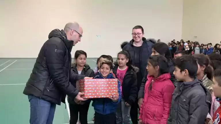 A man in a black jacket hands a wrapped gift box to a smiling boy surrounded by children and adults in a gymnasium. The group looks happy and engaged, with more people gathered in the background.