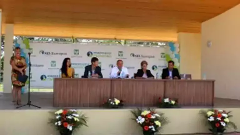 Six people, five seated behind a long wooden table and one standing at a microphone, participate in an outdoor panel discussion on a covered stage decorated with flowers and a branded backdrop.