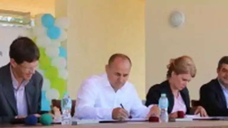 Four people seated at a table during a formal event, with two men and one woman signing documents, and water bottles and microphones on the table. A background banner and balloons are visible.