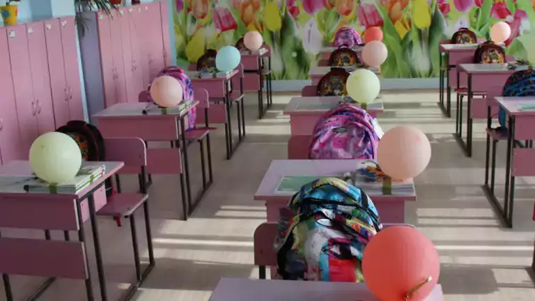 Bright classroom with pink desks and chairs, colorful backpacks, balloons on each desk, and a vibrant tulip flower mural on the back wall. The classroom is well-lit and decorated with plants and books, creating a cheerful and welcoming learning environment.