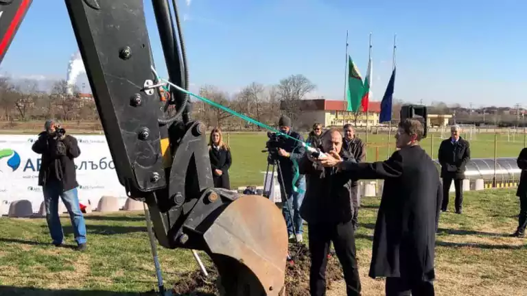 A group of people attends a groundbreaking ceremony outdoors, with two men cutting a ribbon tied to an excavator arm. Flags, a photographer, and a banner are visible in the background on a sunny day.