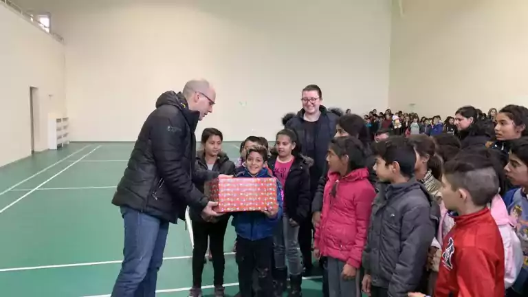 A man in a black jacket hands a wrapped gift box to a smiling boy in a blue coat, surrounded by children and a woman in a gymnasium. The children look happy and engaged, and the gym has a green floor with white lines.