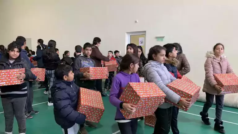 A group of children in winter jackets stand in a gymnasium, each holding a large, red, gift-wrapped box. The children appear to be receiving or distributing presents, and the atmosphere is lively and cheerful.