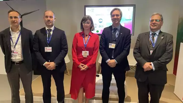 Five professionals, four men in suits and one woman in a red dress, stand side by side in front of a presentation screen at a conference or business event. They are wearing name badges and lanyards, and the setting appears modern and formal.