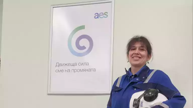A woman in blue work overalls stands indoors holding a white safety helmet. She is smiling and standing next to a wall with an AES company poster and an electronic display showing 167 MW.