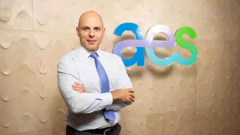 Bald man in a light blue shirt and blue tie stands with arms crossed in front of a beige textured wall and a colorful 'aes' logo.
