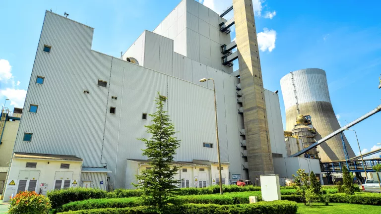 Large industrial power plant with a modern white building, a tall concrete chimney, and a cooling tower in the background, surrounded by green landscaped grass and small trees under a blue sky.