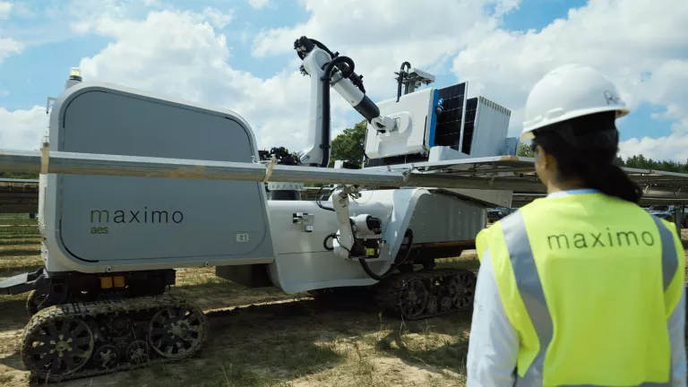 A person in a safety vest and helmet stands in front of a robotic vehicle labeled 'maximo aes' on tracks, inspecting solar panel installations outdoors under a partly cloudy sky.