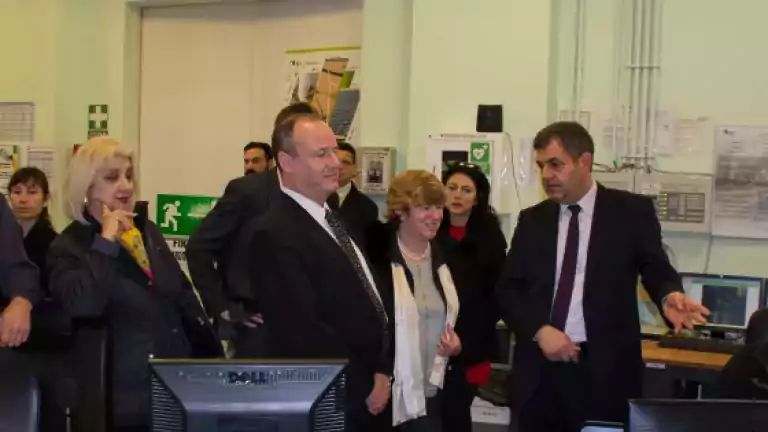 A group of professionally dressed people stands in an office or control room, listening to a man in a suit who is gesturing while speaking. Computer monitors, safety signs, and documents are visible in the background.
