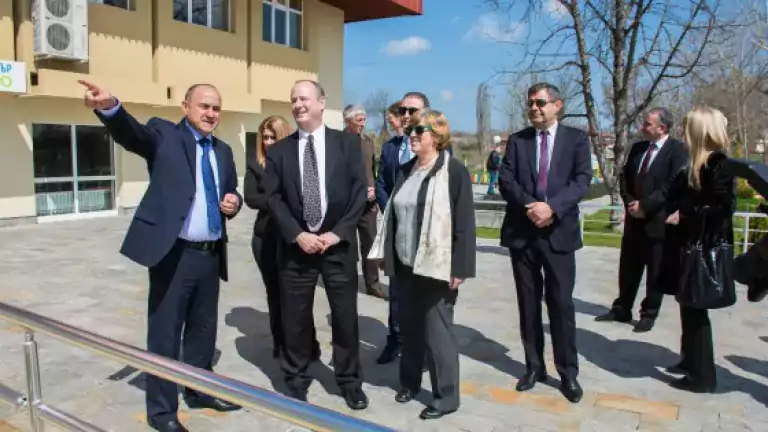 A group of professionally dressed people stands outside a modern building on a sunny day. One man is pointing and appears to be explaining something to the others, who are attentively listening. The scene includes both men and women and a background of trees and blue sky.