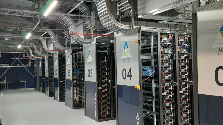 A modern energy storage facility with multiple rows of Advancion battery racks, labeled with numbers and connected by organized wiring and overhead cable trays under bright industrial lighting.