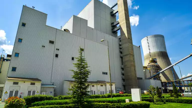 Large industrial power plant with modern white and gray buildings, a tall chimney, and a cooling tower under a blue sky. The foreground features green landscaping with trimmed bushes, small trees, and a grassy lawn.