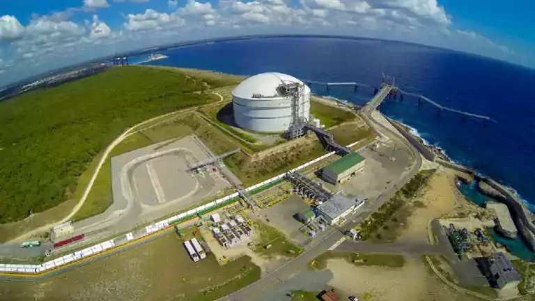 Aerial view of an industrial facility with a large white storage tank near the coast, surrounded by green vegetation, buildings, roads, and a pier extending into the blue ocean under a partly cloudy sky.