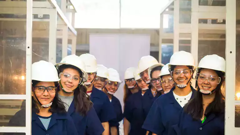 A group of women wearing safety helmets, protective glasses, and blue uniforms stands in two rows inside an industrial or laboratory setting, smiling at the camera.