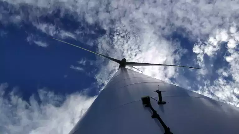 Upward view of a wind turbine against a partly cloudy blue sky, showing the tall white tower and turbine blades extending outward.